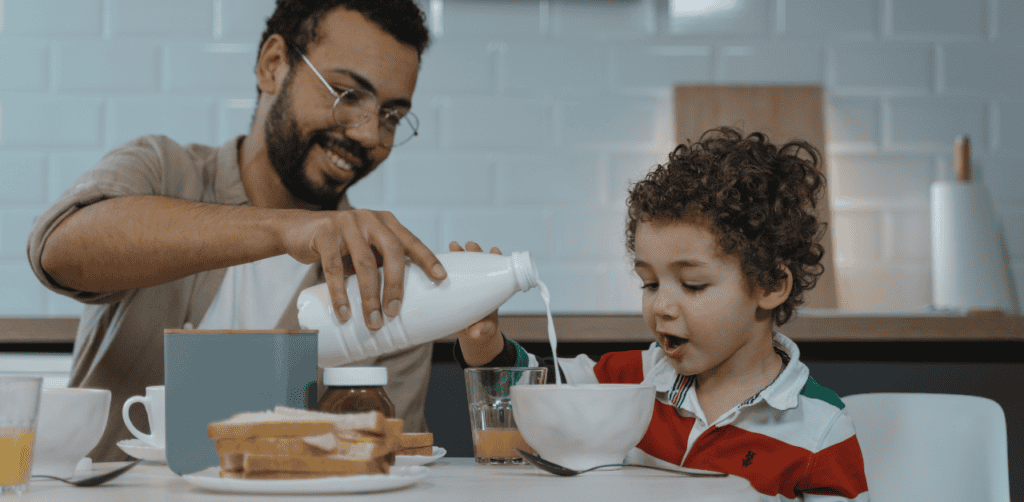 Father and son sharing breakfast, pouring milk into cereal bowl at home.